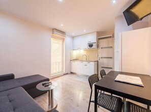 Een knus woongedeelte met een bank, een klein rond tafeltje, een eettafel met stoelen, een kitchenette en een aan de muur bevestigde televisie.