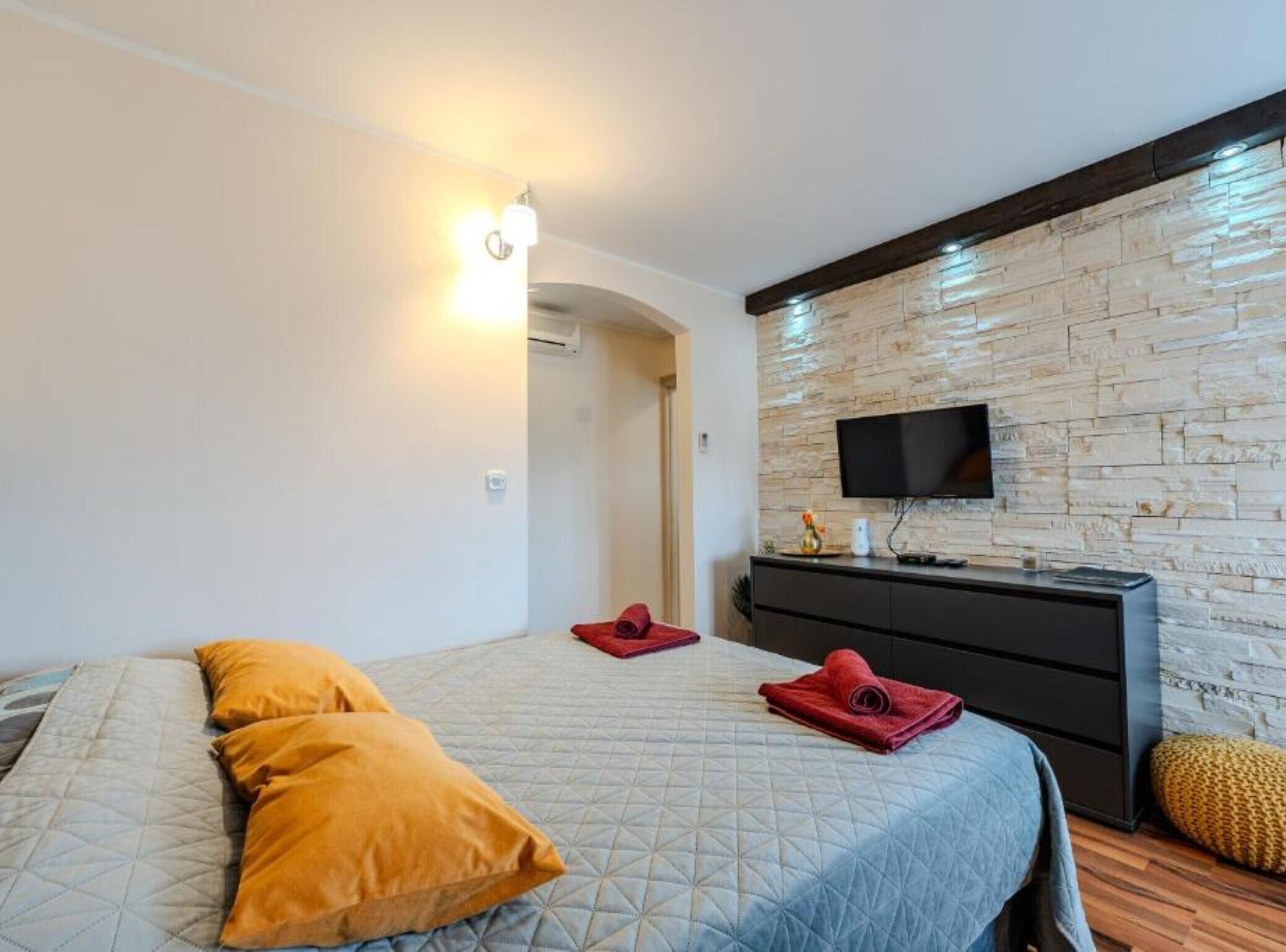 Camera d'albergo con un letto, una cassettiera, una TV montata su una parete in pietra e un corridoio che porta a un'altra stanza.