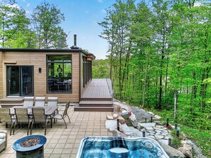 A modern house with a patio, outdoor seating, a hot tub, and a forested backdrop. - Vrbo Property (La Conception)
