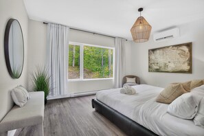 A bedroom with a large window, a bed, a bedside table, a chair, a plant, and a painting on the wall. - Vrbo Property (La Conception)