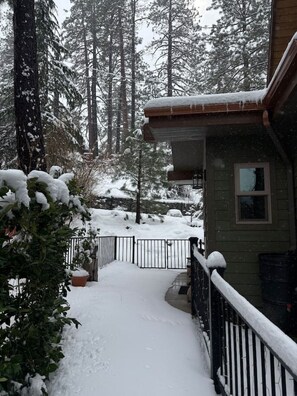 Uma trilha coberta de neve que leva a uma casa com deck de madeira e guarda-corpo de metal.