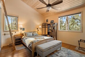 A bedroom with a wooden ceiling fan, a large bed with a patterned bedspread, two bedside lamps, a bookshelf, and a window with a view of trees.