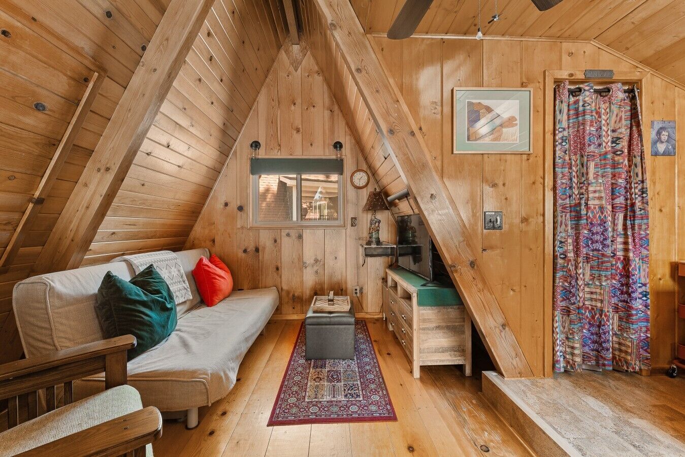 Ett mysigt rum på vinden med ett sluttande tak, en soffa med kuddar, ett litet bord, en mönstrad matta och en dörr med gardiner.