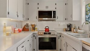 A modern kitchen with white cabinetry, a stainless steel microwave, an oven, and a kettle on the countertop. - Pine & Peak | Cozy Cabin Getaway (Wrightwood)