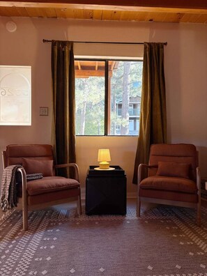 A room with two brown armchairs, a small black side table with a lamp, a patterned rug, a window with curtains, and a wall-mounted light switch. - Pine & Peak | Cozy Cabin Getaway (Wrightwood)