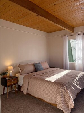 A bedroom with a wooden ceiling, a bed with a checkered bedspread, a bedside table with a lamp, and a window with curtains.