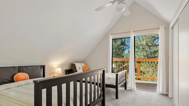 A bedroom with a sloped ceiling, a bed with striped bedding, a nightstand with a lamp, a small table with a chair, and a balcony with a wooden railing. - Pine & Peak | Cozy Cabin Getaway (Wrightwood)