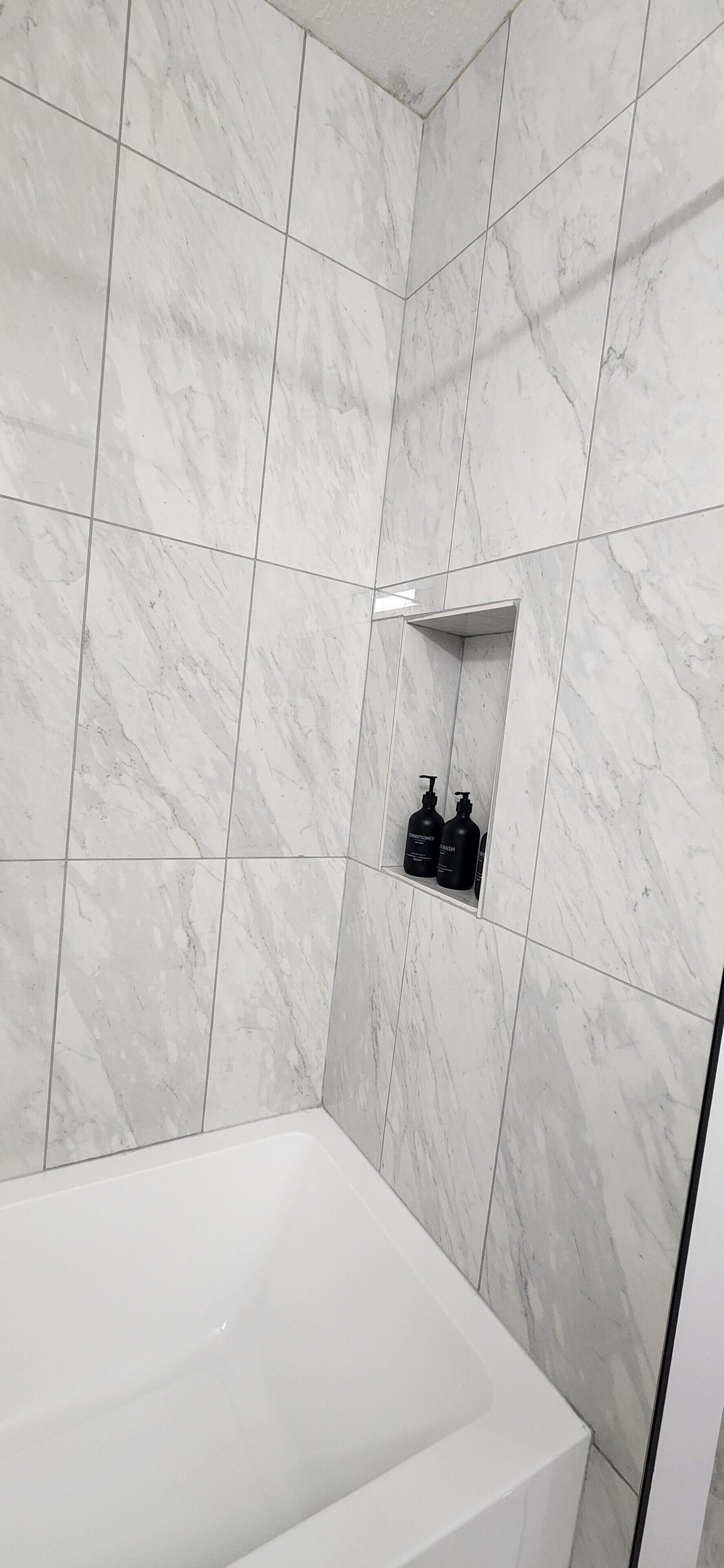 A bathroom with white marble tiles, a white bathtub, and a shelf with two black bottles.