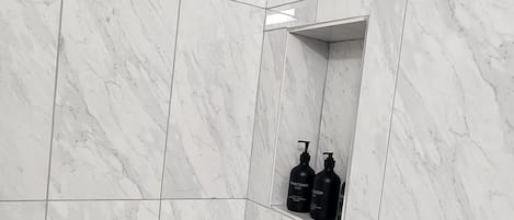 A bathroom with white marble tiles, a white bathtub, and a shelf with two black bottles.