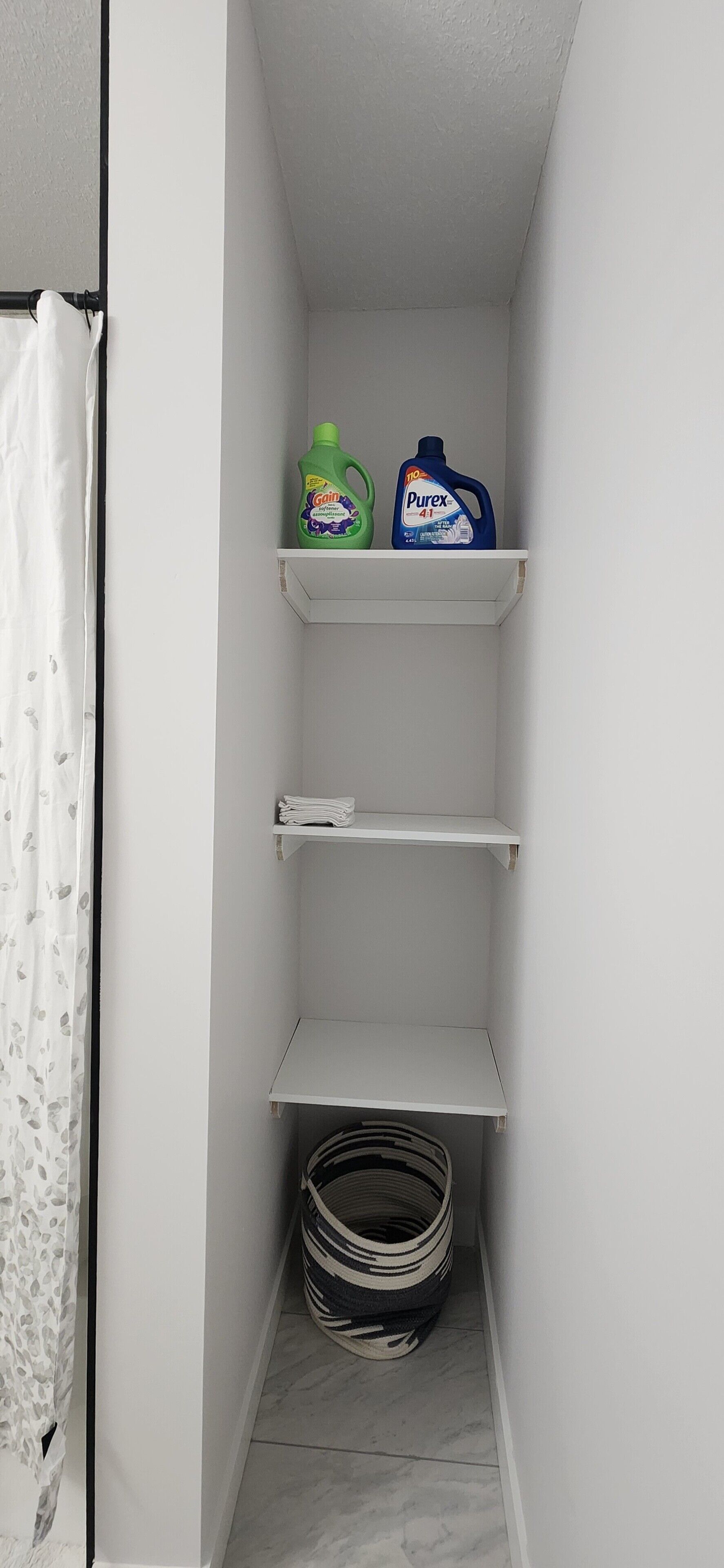 A small laundry room with shelves holding cleaning supplies, a basket, and a curtain.