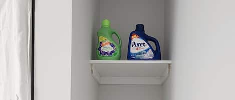 A small laundry room with shelves holding cleaning supplies, a basket, and a curtain.