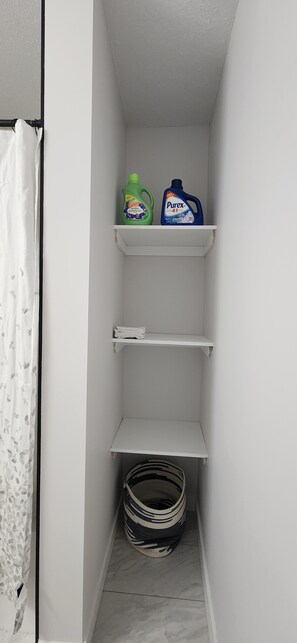 A small laundry room with shelves holding cleaning supplies, a basket, and a curtain. - Modern Cozy One Bedroom suite Near WEM (Edmonton)