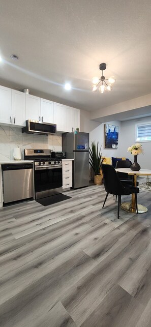 A modern kitchen with white cabinets, stainless steel appliances, and a dining area with a black chair and table. - Modern Cozy One Bedroom suite Near WEM (Edmonton)