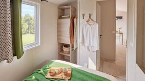 Ett sovrum med en säng, ett fönster med gardiner, en trähylla med kläder och ett bricka med mat.
