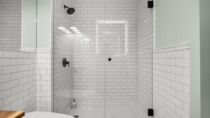 Un baño moderno con azulejos blancos tipo metro, una cabina de ducha de vidrio y un tocador de madera.