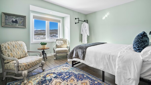 Un dormitorio con una cama, dos sillones, una mesita, una ventana con vistas y un cuadro en la pared.