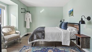 Un dormitorio con cama, una silla, una mesita de noche, una lámpara y un cuadro en la pared.