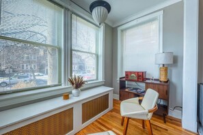 Ett rum med stora fönster, ett element, ett skrivbord med en lampa, en stol och ett elementöverdrag.