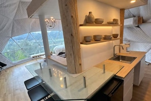 A modern kitchen with a glass dining table, wooden accents, and a view through large windows.