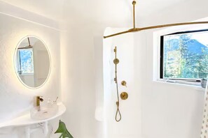 A bathroom with a white sink, a round mirror, a shower with a gold showerhead, and a window with a view of trees.