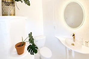 A bathroom with a white sink, a gold faucet, a round mirror, a toilet, and a potted plant.