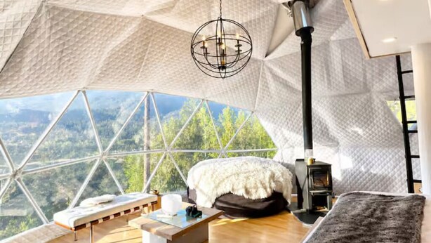 Una habitación tipo cúpula geométrica con una cama, una mesa, una silla, una chimenea y vistas al bosque.