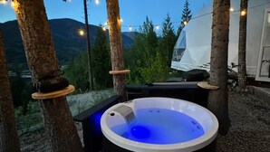 Un jacuzzi con luz azul, rodeado de árboles y una tienda de campaña, con montañas al fondo.