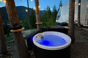 A hot tub with a blue light, surrounded by trees and a tent, with a mountainous background.