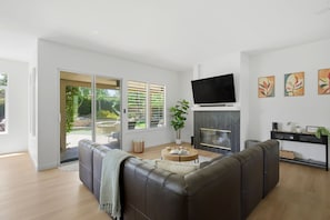 A modern living room with a leather sofa, a flat-screen TV, a fireplace, and a sliding glass door to the outdoors. - Palm Desert Pool Home with Mountain Views (Palm Desert)