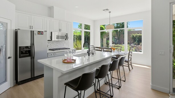 A modern kitchen with white cabinetry, stainless steel appliances, a central island with bar stools, and large windows offering a view of the outdoors. - Palm Desert Pool Home with Mountain Views (Palm Desert)