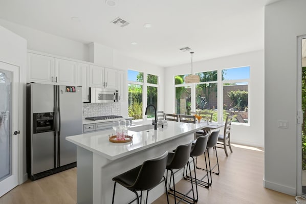A modern kitchen with white cabinetry, stainless steel appliances, a central island with bar stools, and large windows offering a view of the outdoors. - Palm Desert Pool Home with Mountain Views (Palm Desert)