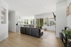 A modern living room with a sectional sofa, a kitchen in the background, and a large window with blinds. - Palm Desert Pool Home with Mountain Views (Palm Desert)