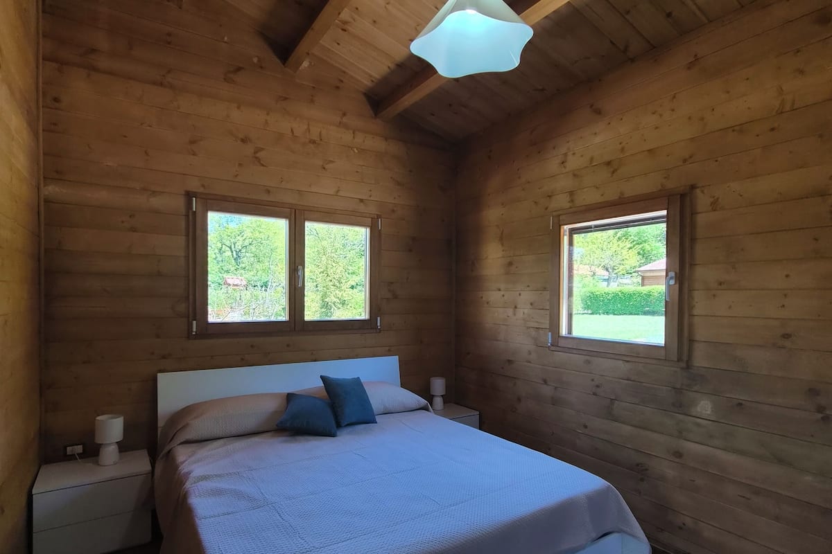 A wooden cabin bedroom with a bed, two windows, a nightstand, and a hanging light fixture.