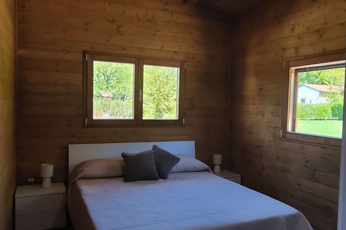 A wooden cabin bedroom with a bed, two windows, a hanging light fixture, and a nightstand with a lamp.