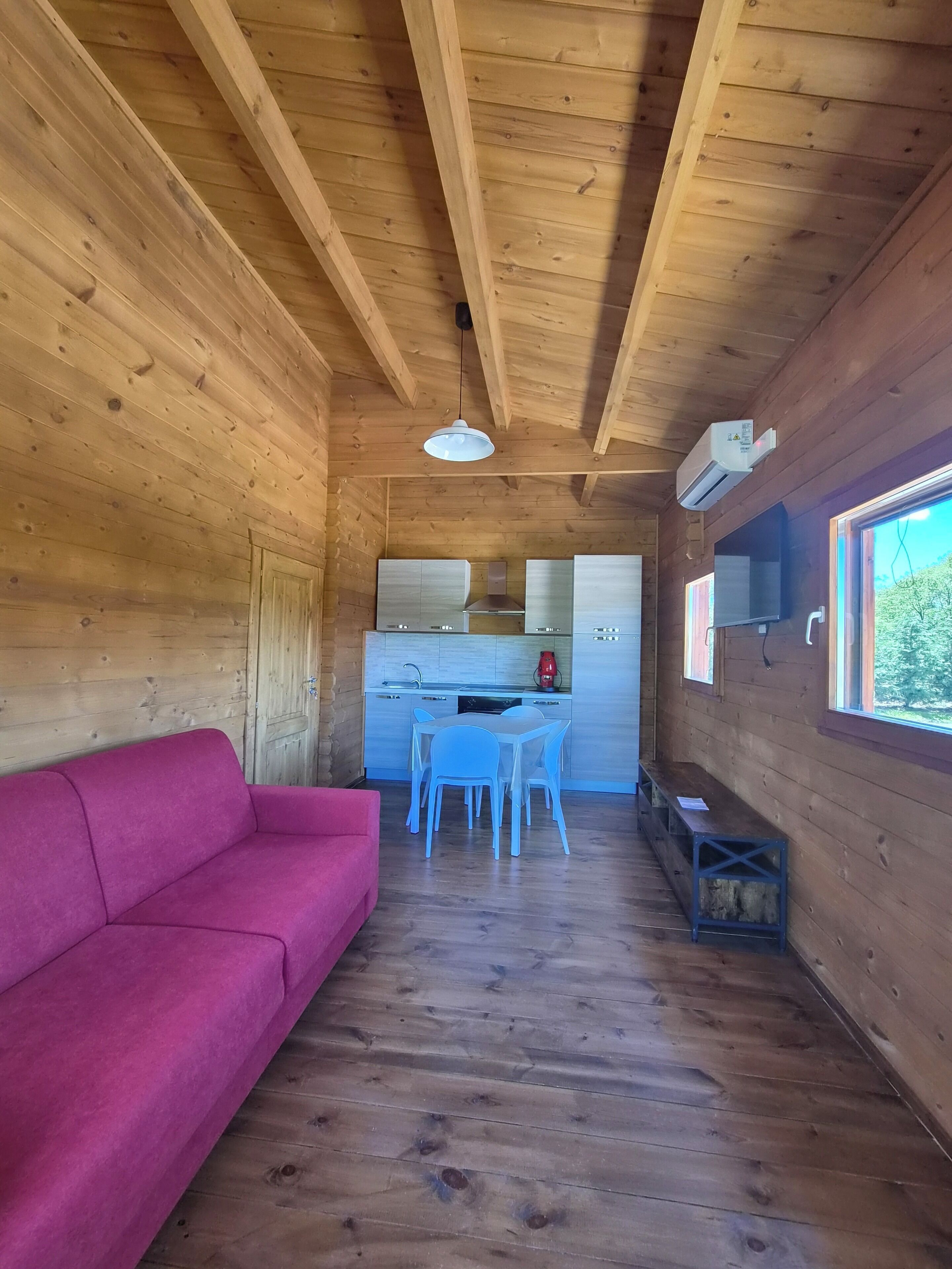 A wooden interior with a sofa, a dining table with chairs, a kitchen area, and a window.