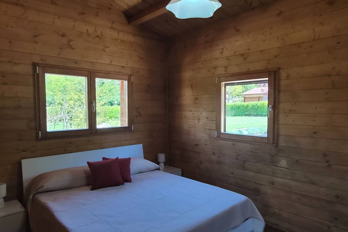 A wooden cabin bedroom with a bed, two windows, a ceiling light, and a nightstand.