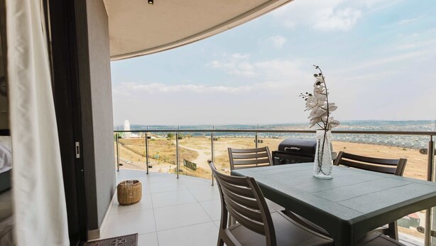 A balcony with a table and chairs, overlooking a beach and a distant cityscape. - The Signature Luxury Stay (Midrand)