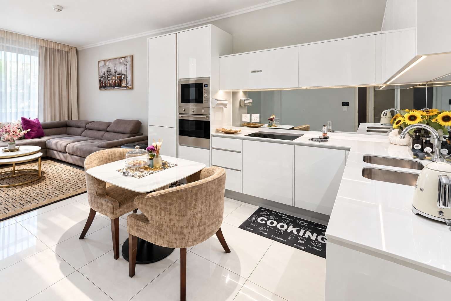 Une cuisine moderne avec des meubles blancs, un coin repas doté d’une table ronde et de chaises, et un salon avec un canapé et un tableau.