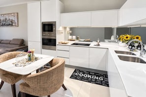 A modern kitchen with white cabinetry, a built-in oven, a microwave, and a sink. There is a dining area with a table and chairs, and a rug with the word 'COOKING' on it.