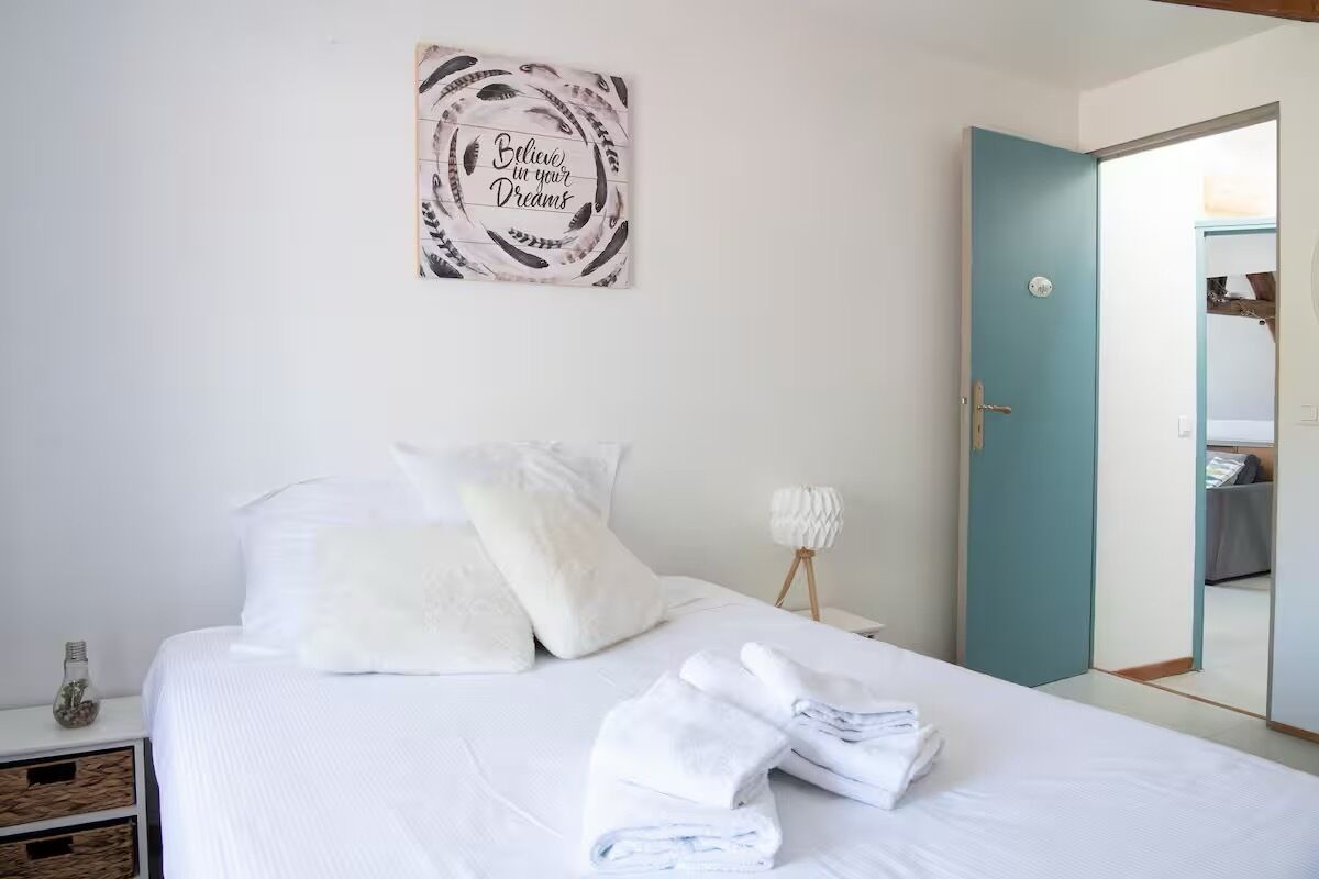 A bedroom with a bed, pillows, a folded towel, a wall art piece, a bedside lamp, and a door leading to another room.