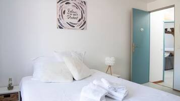 A bedroom with a bed, pillows, a folded towel, a wall art piece, a bedside lamp, and a door leading to another room.