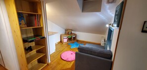 Ein Zimmer mit einem Holzbücherregal, einem kleinen Tisch mit einem Spielzeug, einer rosa Matte und einem grauen Sofa.