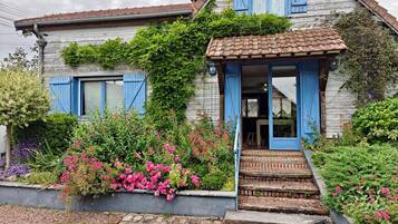 A rustic house with blue shutters, a tiled roof, and a front garden with pink flowers.