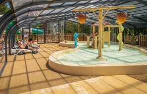 Un espace de jeux aquatiques couvert, dotĂ© de toboggans et dâune piscine, entourĂ© dâune terrasse en bois.