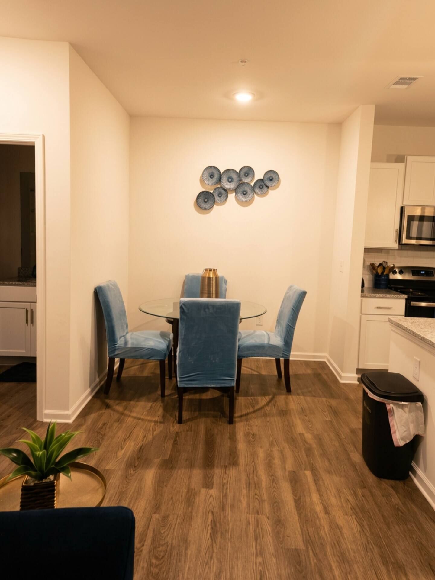 A dining area with a glass table and blue chairs, a kitchen with white cabinets, and a wooden floor.