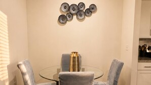 Een eethoek met een glazen tafel, omringd door vier stoelen, een decoratief kunstwerk erboven en een keuken aan de rechterkant.