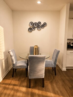 A dining area with a glass table surrounded by four chairs, a decorative wall piece above the table, and a kitchen area to the right. - King Bed Suite, 8 Min Airport, Fast WiFi & Gym (Jacksonville)