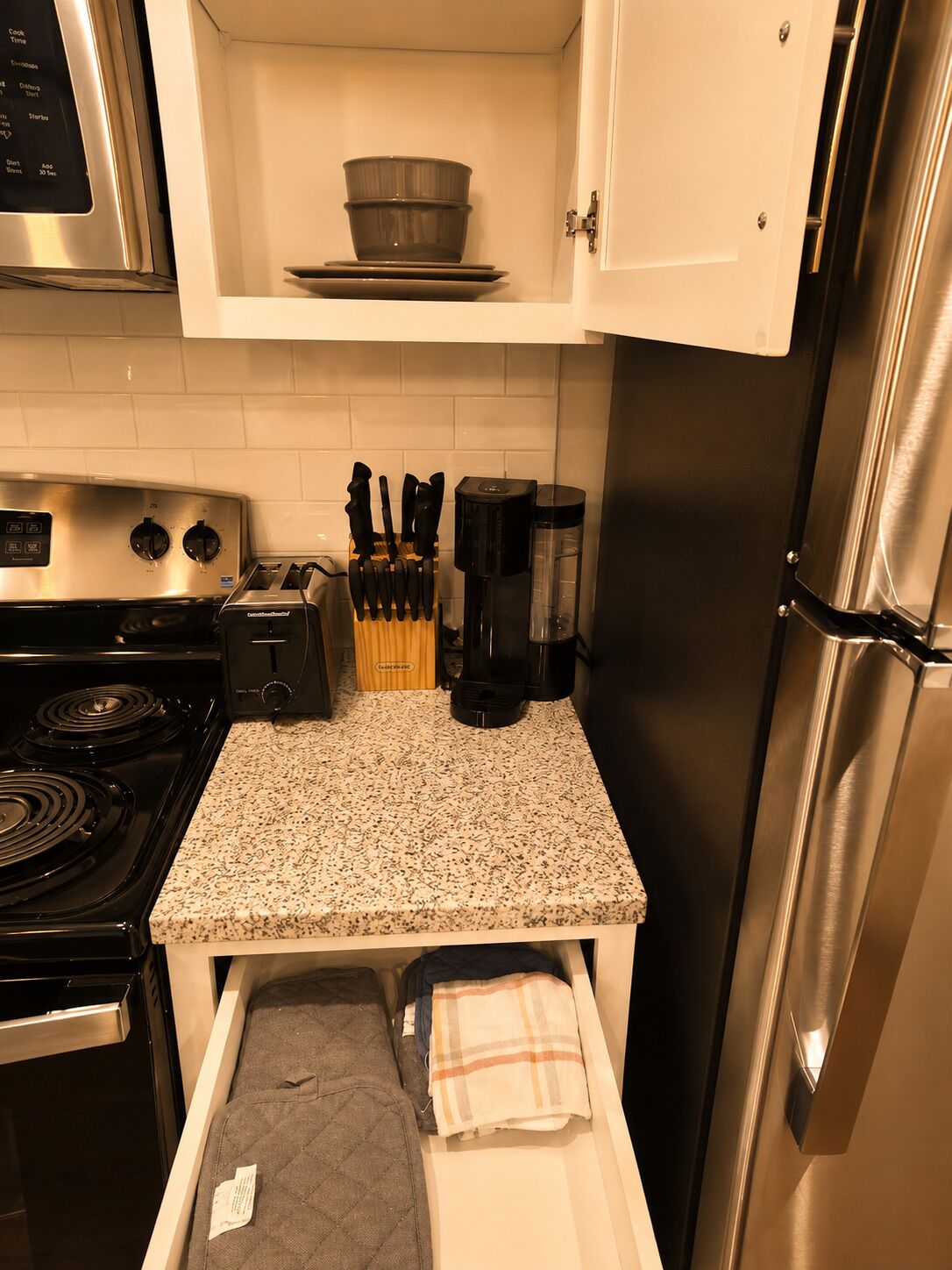 A compact kitchen with a stove, toaster, coffee maker, and a drawer with a towel.