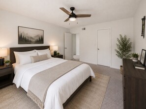 Une chambre à coucher avec un lit, des tables de chevet, un ventilateur de plafond, un tableau, une porte et une plante.