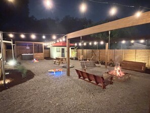 Ein Terrassenbereich im Freien mit einem Holztisch, Bänken, einer Feuerstelle und Lichterketten.